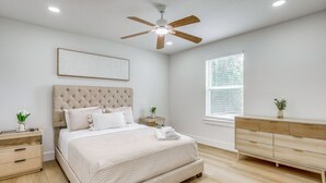 Quarto com uma cama grande, um ventilador de teto, uma mesa de cabeceira com uma planta e uma janela com persianas.