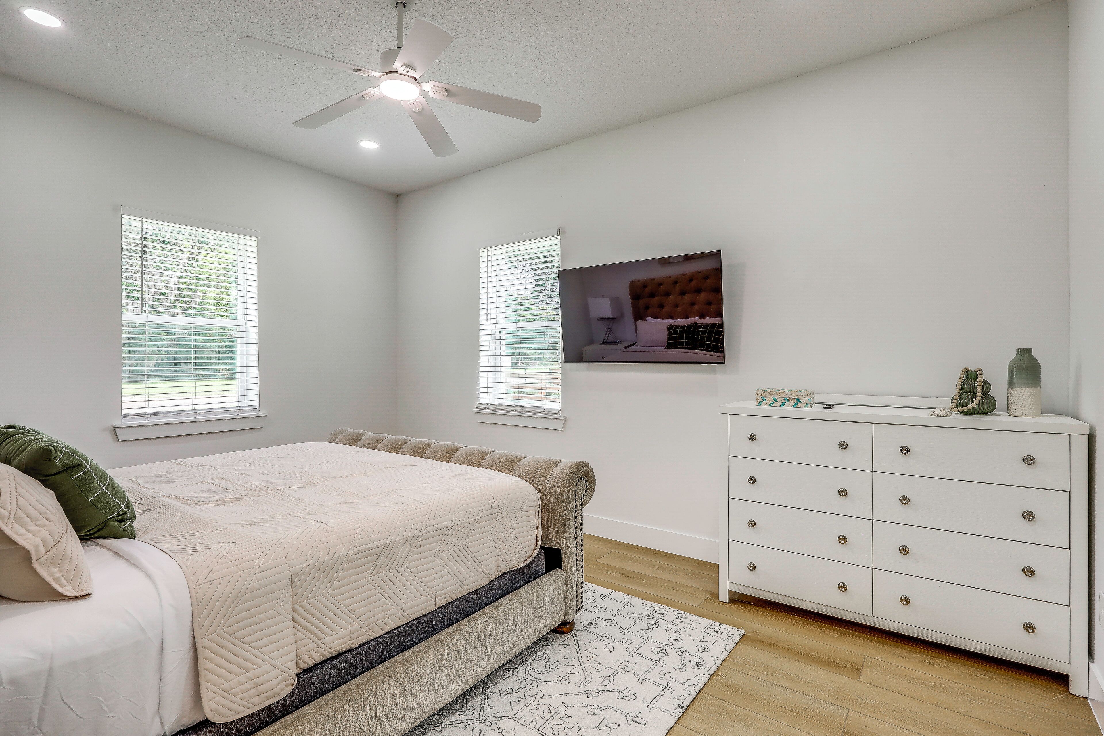 Ein Schlafzimmer mit Bett, Schminktisch, Deckenventilator und zwei Fenstern mit Jalousien.