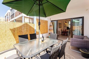 Une table en verre avec des chaises, un parasol vert, un canapé en osier et un mur en bambou.