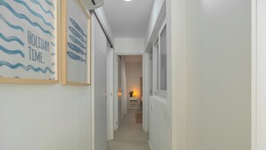 Um corredor estreito com um quadro na parede esquerda, uma mesinha com uma planta no final e uma janela à direita.