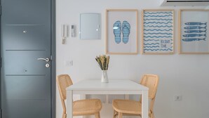 Una habitación con puerta gris, paredes blancas, una mesa blanca con dos sillas de mimbre, un jarrón con flores secas y tres cuadros en la pared.