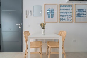 Ein Zimmer mit einer grauen Tür, weißen Wänden, einem weißen Tisch mit zwei Korbstühlen, einer Vase mit getrockneten Blumen und drei gerahmten Bildern an der Wand.