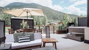 Eine Außenpatio mit einer Couch, einem Tisch und einem Sonnenschirm, umgeben von Blumentöpfen und Blick auf einen bewaldeten Berg.