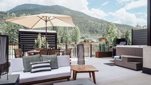 Eine Außenpatio mit einer Couch, einem Tisch und einem Sonnenschirm, umgeben von Blumentöpfen und Blick auf einen bewaldeten Berg.
