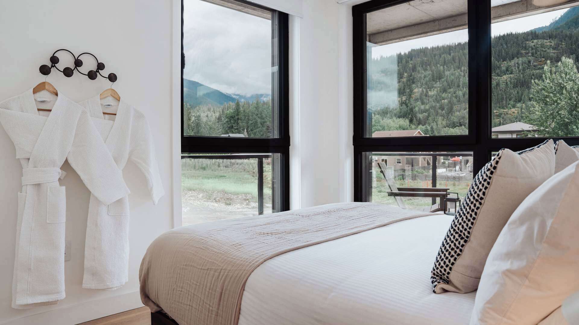 A bedroom with a large bed, two white robes hanging on a rack, and a view of a forested mountain area through large windows.