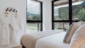Ein Schlafzimmer mit einem großen Bett, zwei weiße Bademäntel an einem Gestell und Blick auf eine bewaldete Berglandschaft durch große Fenster.
