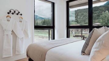 A bedroom with a large bed, two white robes hanging on a rack, and a view of a forested mountain area through large windows.