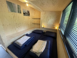 Een houten kamer met twee bedden, een plank met verlichting en een raam met jaloezieën.