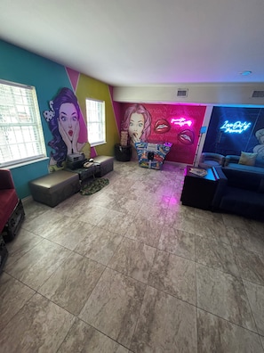 A room with a large mural, arcade machines, a neon sign, and a sofa.