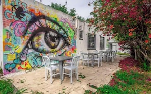 Ein Außenbereich mit Tischen und Stühlen, einem Wandbild mit Graffiti und einem Gebäude mit Fenstern.