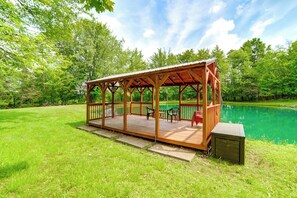 Een houten gazebo met terras, omringd door een gazon en een vijver.