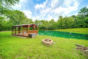 Eine hölzerne Laube mit einer Terrasse, einem Feuerkorb und einem Teich in einer grünen Wiese.