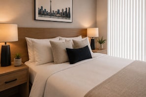 A hotel room with a bed, two bedside tables, a nightstand, a framed cityscape picture, and a window with blinds. - Norvian Urban Suites (Toronto)