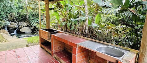 A covered outdoor area with a brick-built sink and bench, surrounded by lush greenery and a small waterfall.