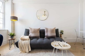Ein modernes Wohnzimmer mit einer Couch, einem runden Spiegel, einer Stehlampe und einem kleinen Tisch mit Gläsern.