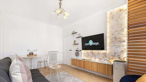 A modern living room with a sofa, a coffee table, a TV mounted on a marble wall, and a small kitchen area with a refrigerator. - St John's Wood Flat with Spa Bath (Greater London)