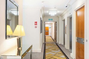 Un couloir d’hôtel avec un tapis à motifs, un panneau indiquant la sortie de secours et un miroir sur le côté gauche.