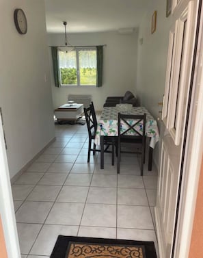A narrow hallway leading to a dining area with a table and chairs, a radiator, and a window with curtains.