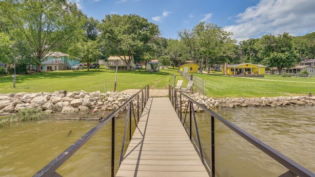 Eine Holzbrücke führt zu einer Wiese mit Häusern und einem Teich.