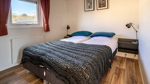 A bedroom with a large window, a bed with a knitted blanket, two pillows, a bedside table with a lamp, and two framed pictures on the wall. - Velthorst Waterfront 6 (Linden)