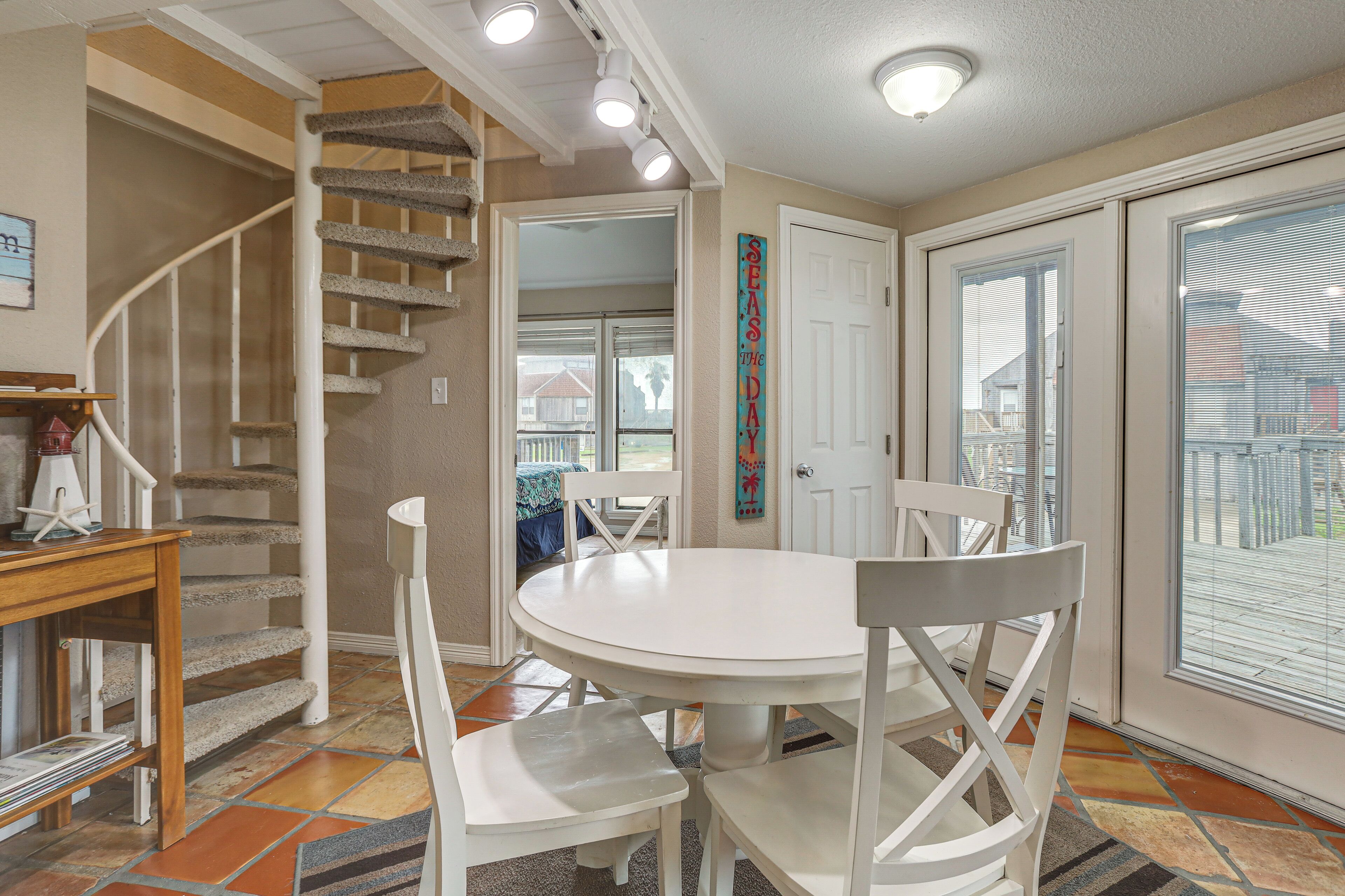 Un coin repas avec une table ronde, des chaises blanches, un escalier et une vue à travers une porte vitrée.