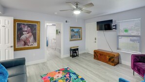 Una sala de estar con ventilador de techo, televisor montado en la pared, un cofre de madera y un tapete colorido.