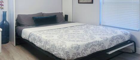 A bedroom with a bed, a nightstand, a wall-mounted picture, and a window with blinds.