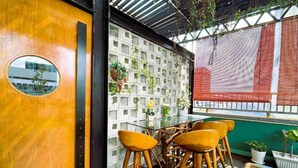 A room with a wooden door, a round mirror, a glass table with wooden chairs, a white lattice wall with plants, and a view through a window with a red blind. - Enclave Hotel Kuala Lumpur (Kuala Lumpur)