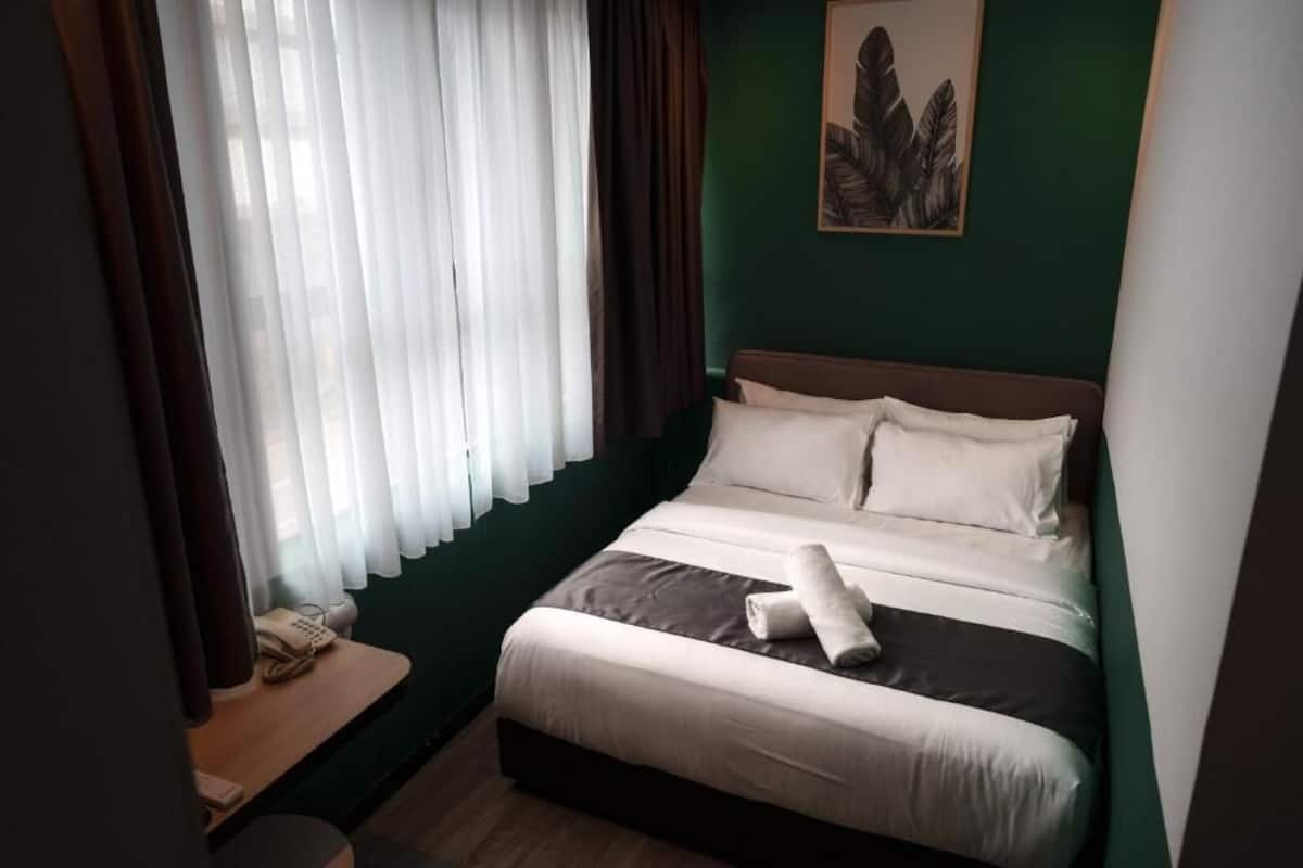 Ein ordentlich bezogenes Bett mit Kissen, ein Nachttisch mit Telefon und ein Fenster mit durchsichtigen Vorhängen.