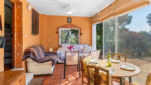 A sunlit room with a sofa, a dining table, and a window overlooking a garden. - Estepona Mirador Country House (9 km from the beach) (Málaga)