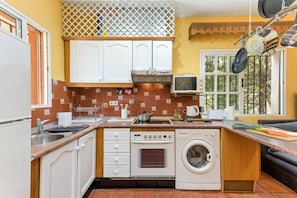 A compact kitchen with white cabinets, a built-in oven, a washing machine, and a window with hanging pots.