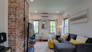 A living room with a brick wall, a gray sofa, a yellow ottoman, a white armchair, and a large window. - Extended East Knoxville Stay, 2 Mi to Dtwn! (Knoxville)