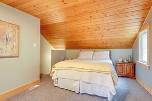 Une chambre avec un plafond en bois incliné, un lit recouvert de plusieurs couches de literie, une table de chevet agrémentée d’objets décoratifs et une fenêtre laissant entrer la lumière naturelle.