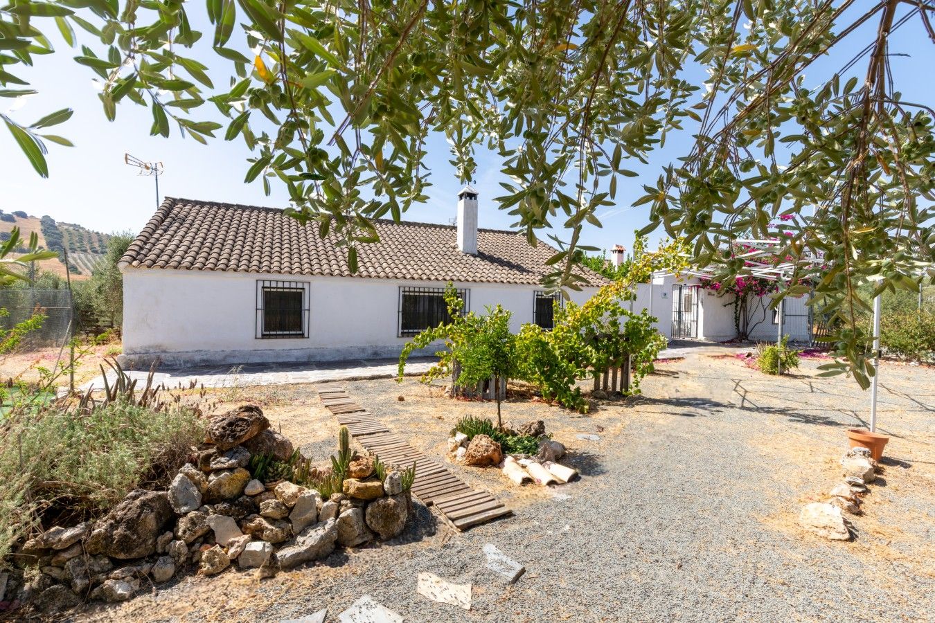 A white building with a tiled roof, surrounded by a gravel path, small trees, and a stone wall.