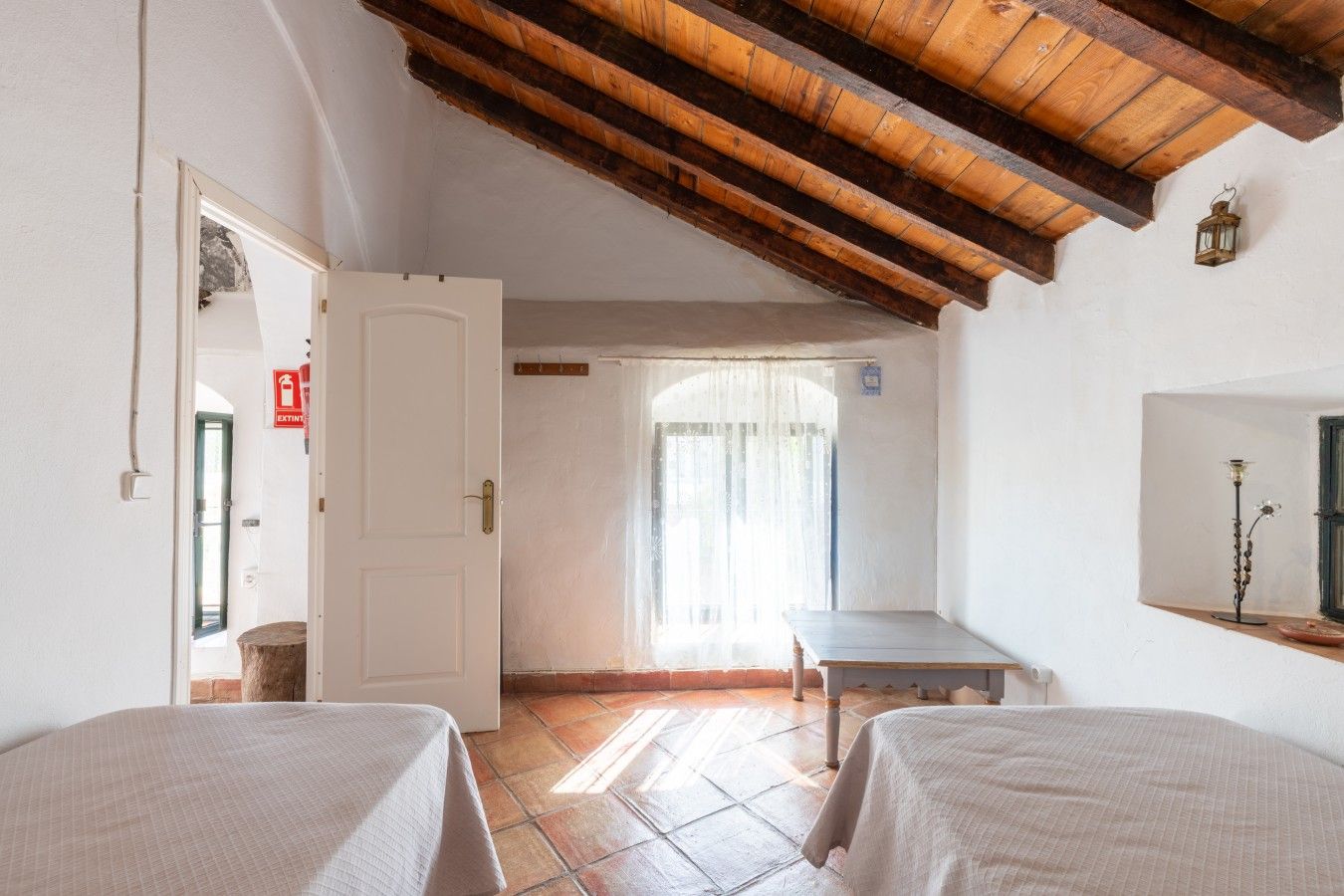 Una habitación con dos camas, techo de madera, ventana con cortinas y una pequeña mesa.
