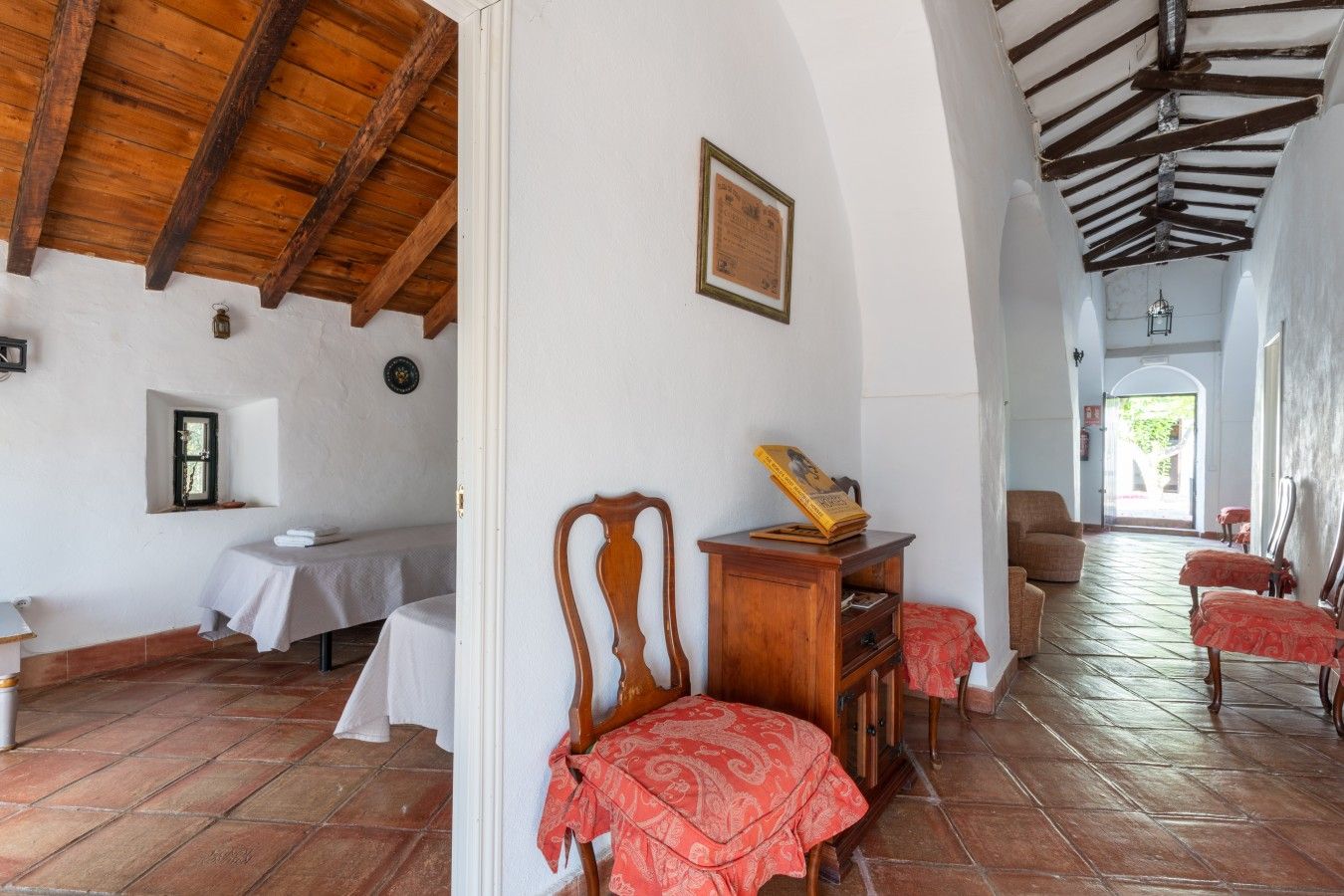 A room with a bed, a wooden chair, a table, and a red patterned tablecloth.