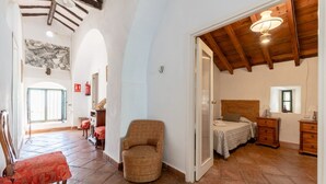Una habitación amplia con una cama, una silla y una mesita. Hay una puerta que da a otra habitación.