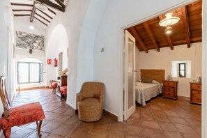 Una habitació àmplia amb un llit, una cadira i una taula petita. Hi ha una porta que dona a una altra habitació.