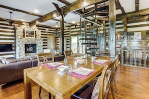 A rustic cabin interior with a dining table set for four, a stone fireplace, wooden beams, and a staircase.