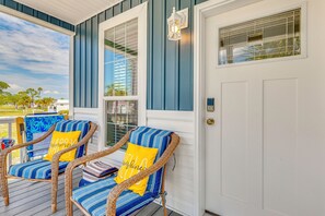 Eine Veranda mit zwei Korbstühlen, einer weißen Tür und einem Fenster mit Blick auf Palmen.