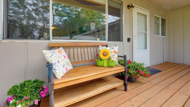 Un banc en bois avec un coussin, un tournesol en pot et une plante en pot aux fleurs roses, le tout sur un porche.