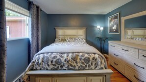 Une chambre à coucher comprenant un lit, une commode, une table de chevet, une fenêtre avec des rideaux et une lampe fixée au mur.