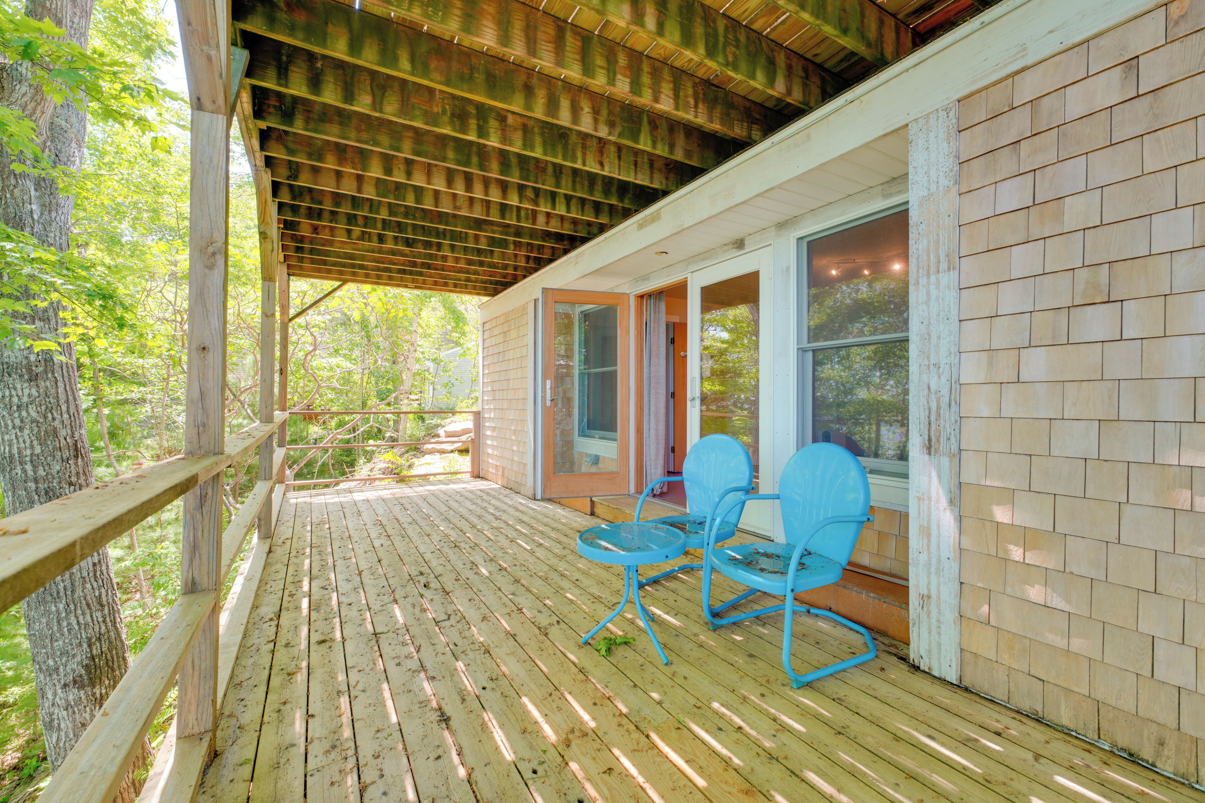 A covered wooden deck with two blue chairs and a table, a wooden railing, and a brick wall.