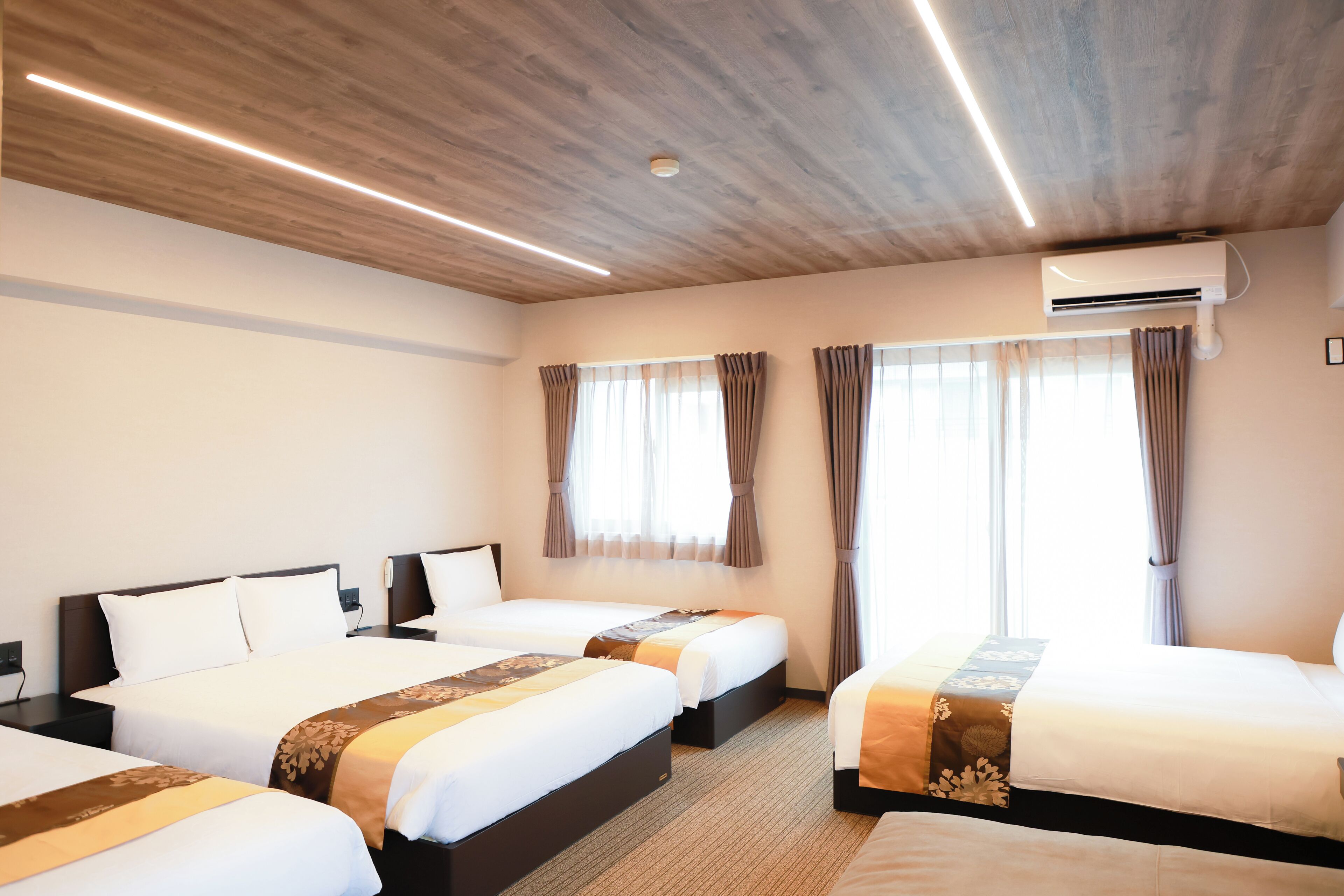 A hotel room with three beds, wooden ceiling, air conditioning unit, and large windows with curtains.