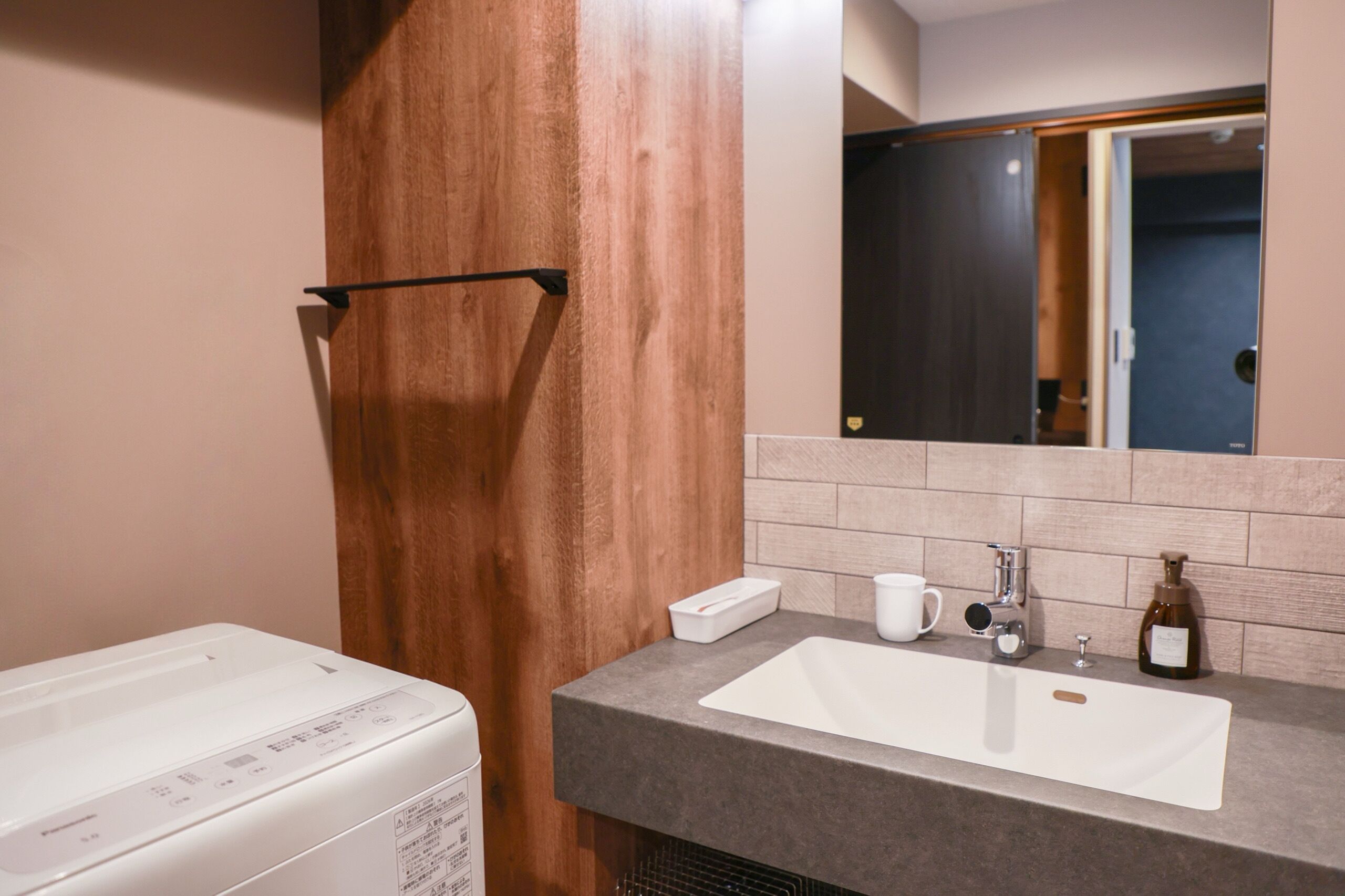 A bathroom with a wooden door, a white washing machine, a sink with a mirror above it, and a bottle of soap.