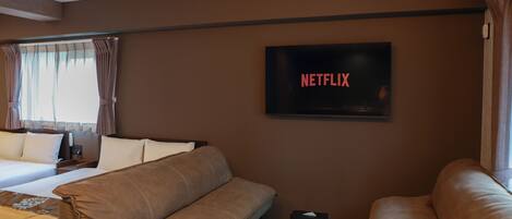 A hotel room with a bed, two sofas, a TV displaying 'NETFLIX,' and a window with curtains.