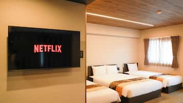 A hotel room with a television displaying 'NETFLIX,' two beds with white bedding, a wooden ceiling, and a window with curtains.