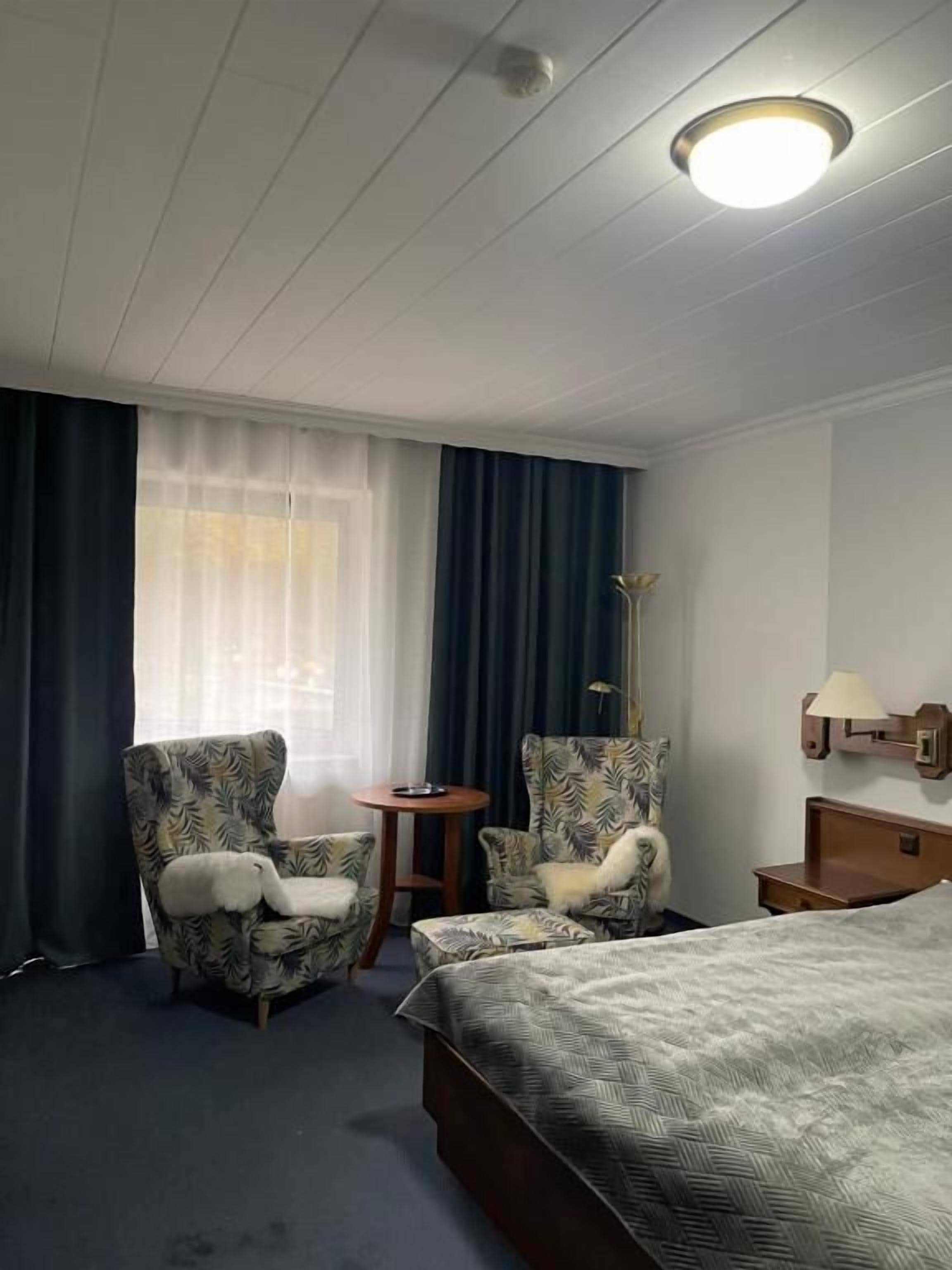 Une chambre d’hôtel avec un lit, deux fauteuils, une petite table, une lampe et une fenêtre avec des rideaux.