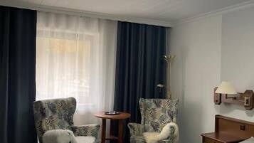 Une chambre d’hôtel avec un lit, deux fauteuils, une petite table, une lampe et une fenêtre avec des rideaux.