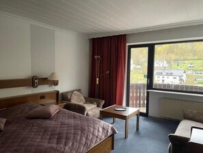 Habitació d'hotel amb llit, cadira, taula, làmpada i vista d'un edifici a través d'una finestra gran.